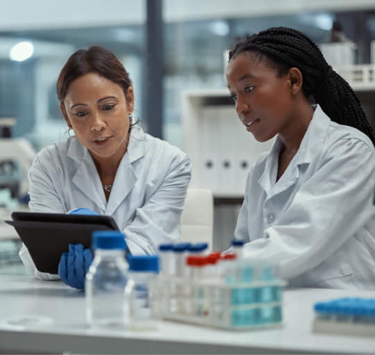 Two women in lab coats looking at a tablet, with vials sitting on the table in front of them